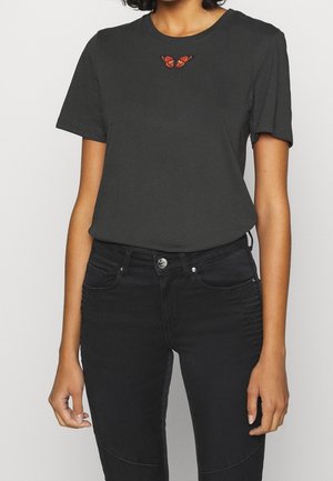 Person wearing black T-shirt with small orange butterfly design at neckline and black fitted pants, standing against a plain background.