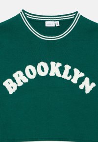 Zeleni pulover z rebrastim belim ovratnikom, na katerem so puhasti beli črki "BROOKLYN" čez sprednji del. Mehka pletenina.