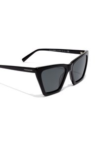 Black plastic sunglasses with a geometric cat-eye shape, dark lenses, and engraved branding on the temples. Smooth texture and minimalist design.