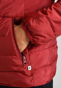 Ruka vložená do zipové kapsy červené prošívané bundy s elastickým manžetem a malou textilní štítkovou nášivkou.