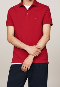 Homme portant un polo rouge à manches courtes avec un petit logo sur la poitrine, un pantalon bleu marine et un t-shirt blanc visible au col et à l'ourlet.