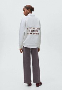 Una donna con i capelli castani raccolti in un bun indossa una felpa grigia chiara con la scritta "LA MIA PLAYLIST È MEGLIO DELLA TUA" sul retro, pantaloni scuri e scarpe bordeaux.