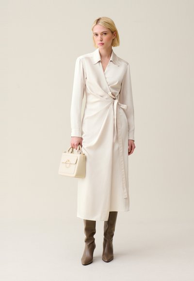 Robe enveloppante couleur crème avec des manches longues, un lien à la taille et une silhouette fluide, associée à des bottes taupe jusqu'aux genoux et un petit sac à main crème.