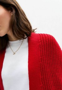 Röd stickad cardigan med ett strukturerat mönster, öppen front och långa ärmar, bärs över en vit t-shirt, med ett guldhalsband.