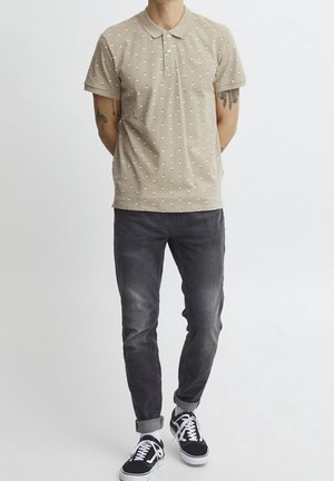 Polo beige avec un motif texturé de petits diamants blancs et un col. Porté avec un jean gris et des baskets noires.