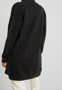 Tmavě šedý pletený cardigan s uvolněným střihem, dlouhými rukávy a designem s asymetrickým lemem. Měkká textura s viditelným minimálním zdobením.