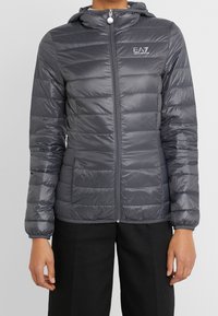 Chaqueta acolchada en gris oscuro, con capucha, cierre de cremallera y diseño acolchado horizontal. El material parece brillante y ligero.