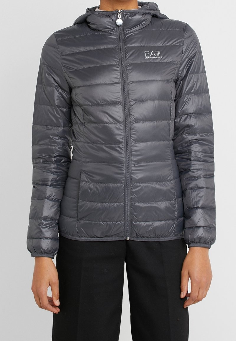 Chaqueta acolchada en gris oscuro, con capucha, cierre de cremallera y diseño acolchado horizontal. El material parece brillante y ligero.