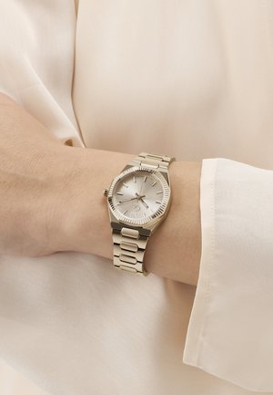 Zilveren polshorloge met metalen band om de linkerpols, gedragen over een beige shirt met lange mouwen, wijzend op ongeveer 10:09 uur.