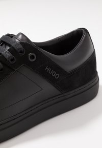 Musta lenkkari, jossa on yhdistelmä sileää nahkaa ja mokkaa, pyöreä kärki, mustat nauhat ja logotieto sivussa.