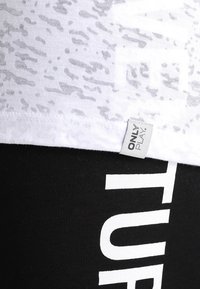 Biały sportowy top z subtelnym wzorem, z metką oznaczoną "ONLY PLAY." Czarne legginsy z wyraźnym białym napisem na nodze.