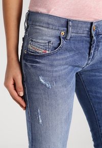 Denim-Jeans in einem Farbverlauf von Blau, mit einem ausgefransten Look und stofflichen Abschnitten sowie einem Patch-Label auf der vorderen Tasche.