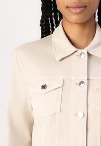 Beige denim jacket featuring a collar, two chest pockets with black hardware, and silver buttons. The fabric has a smooth texture.