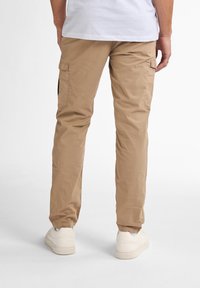 Beige cargo bukser med to sidelommer, en afslappet pasform og elastiske manchetter. Bæres med hvide sneakers og en almindelig hvid t-shirt.
