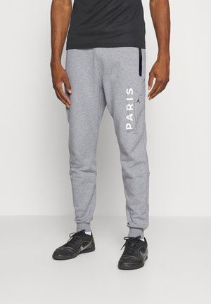 Mand iført lysegrå joggingbukser med teksten "PARIS" på venstre ben og sorte sneakers, stående mod en ensfarvet lys baggrund.
