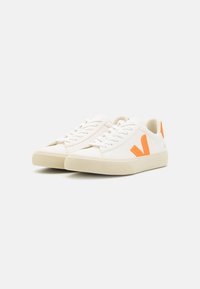 Sneakers bianchi in tela con accenti arancioni in gomma, caratterizzati da una suola testurizzata e un tradizionale sistema di allacciatura. Design semplice e moderno.