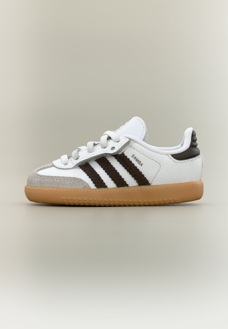 Λευκό αθλητικό παπούτσι Adidas Samba με μαύρες ρίγες, καουτσούκ σόλα, γκρι δερμάτινο πατούρα και λευκά κορδόνια, εμφανίζεται από την εξωτερική πλευρά.