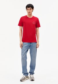 T-shirt en coton rouge à manches courtes, col rond, avec un petit imprimé circulaire sur la poitrine. Associé à un jean en denim bleu clair.