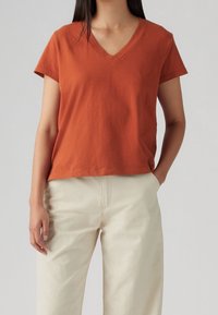 T-shirt à col en V court en coton doux de couleur rouille, associé à un pantalon ample de couleur crème.