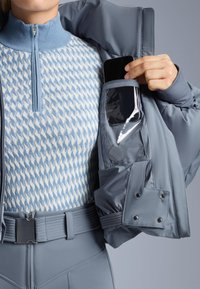 Svijetlosivi sako s džepom na zatvarač u kojem se nalazi pametni telefon, nosi se preko uzorčane svijetloplave pletive majice s visokim ovratnikom i uskim remenom.