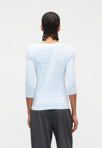 Άτομο με κοτσίδα που φοράει πουκάμισο με ριγέ τρία τέταρτα μανίκια σε ανοιχτό μπλε και λευκό και σκούρο γκρι παντελόνι, με την πλάτη γυρισμένη.