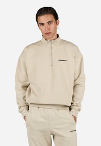 Beige zip-up hettegenser i myk stoff med høy krage og avslappet passform. Har en svart "PEGADOR" logo på brystet.