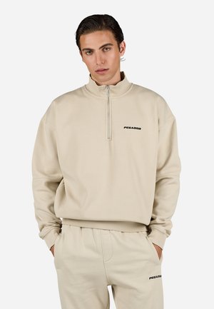 Beige zip-up hettegenser i myk stoff med høy krage og avslappet passform. Har en svart "PEGADOR" logo på brystet.