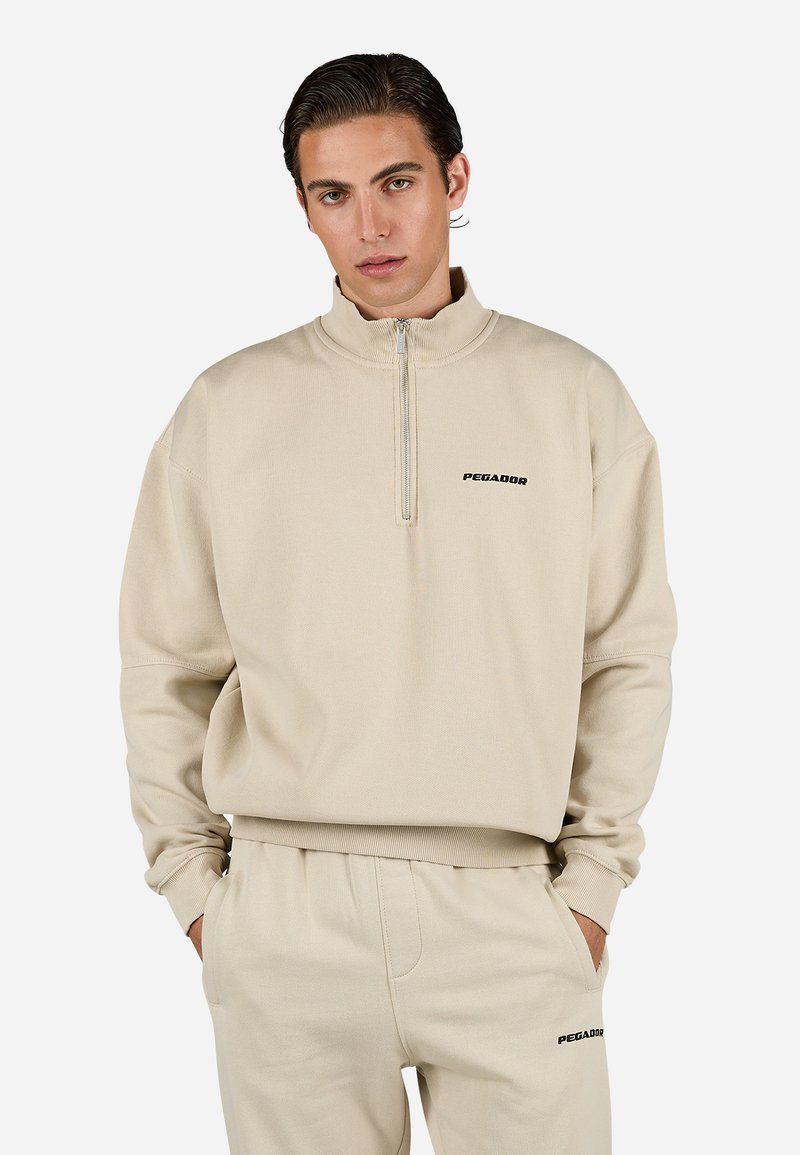 Beige zip-up hettegenser i myk stoff med høy krage og avslappet passform. Har en svart "PEGADOR" logo på brystet.