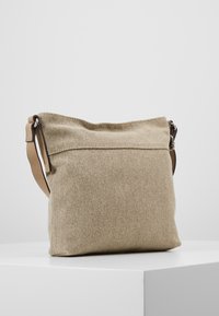 Beige textilaxelväska med en mjuk, slapp design. Har en brun läderrem och en vikt topp för enkel åtkomst. Mjuk textur.