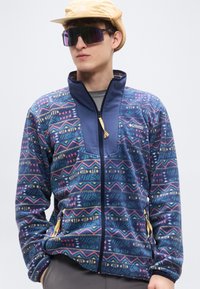 Ζακέτα από φλις με πολύχρωμο γεωμετρικό σχέδιο σε ναυτικό μπλε βάση, με ψηλό γιακά και τσέπες με φερμουάρ για πρόσθετη λειτουργικότητα.