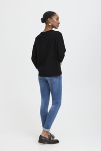 Žena s tmavými vlasy v drdolu, oblečená ve zlatých kroužkových naušnicích, černém dlouhém rukávu, s ohrnutými modrými skinny džíny a tmavými mokasíny, stojí bokem.