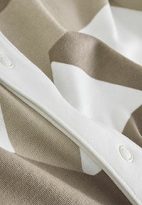 Tissu en coton présentant un motif géométrique beige et blanc, avec une bordure blanche et des fermetures à pression pour un détail supplémentaire. Texture douce.