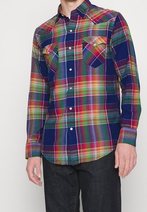 Mężczyzna ubrany w wielokolorową koszulę w kratę z długim rękawem, z dwoma kieszeniami na piersi, oraz ciemne spodnie.