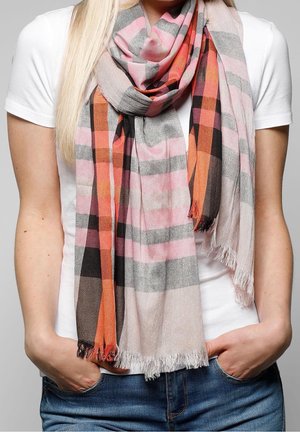 Personne portant une écharpe à carreaux multicolore en rose, orange, noir et gris sur une chemise blanche, les mains dans les poches d'un jean bleu.