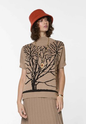 Vrouw draagt een beige kortemouwen trui met zwarte boombedrukking, bijpassende geplooide rok, rode hoed, gouden ketting en armband.