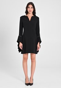 Schwarzes Kleid mit V-Ausschnitt, ausgestellten Ärmeln und Rüsche am Saum. Hergestellt aus strukturiertem Stoff mit dezenten silbernen Akzenten an den Ärmeln.