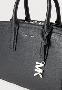 Sac à main en cuir noir avec une surface texturée, des ferrures en argent et un charm emblématique MK attaché à la tirette de la fermeture.