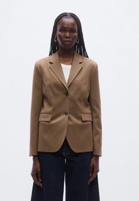 Brun skræddersyet blazer med to knapper, indsnørede revers og sidelommer. Stoffet har en glat tekstur. Båret over en hvid top.