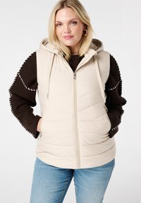 Beige gewatteerde vest met capuchon, voorzien van een gestikt ontwerp en ritssluiting. Draagt over een donkerbruin sweater en blauwe jeans.