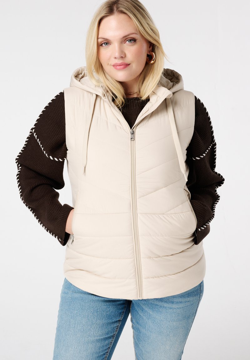 Beige gewatteerde vest met capuchon, voorzien van een gestikt ontwerp en ritssluiting. Draagt over een donkerbruin sweater en blauwe jeans.