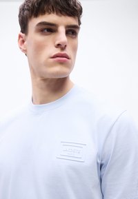 Νεαρός άνδρας με κοντά καστανά μαλλιά κοιτά δεξιά, φορώντας ένα ανοιχτό μπλε T-shirt Lacoste Paris σε λευκό απλό φόντο.