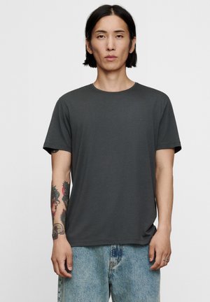 Jong persoon met halflang zwart haar, draag een donkergrijs T-shirt en lichtblauwe jeans, met kleurrijke tatoeages op de linker onderarm.