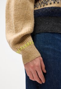 Ullgenser med en beige kropp, grå ribbestrikket kant og ermer med grønne aksentsting. Nærbilde av ermet og hånden, som viser tekstur.