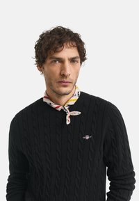 Pulover negru din lână tricotat cu model în cablu, având o textură cu nervuri, prezentând un mic logo brodat. Accesorizat cu o eșarfă la gât cu un model multicolor.