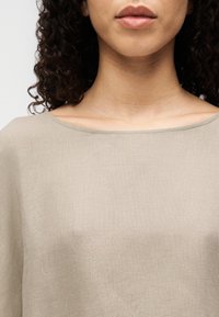 Bež bombažna zgornja majica s širokim izrezom in kratkimi rokavi. Tkanina ima teksturiran, rahlo mat videz. Pod izrezom je vidna koža.