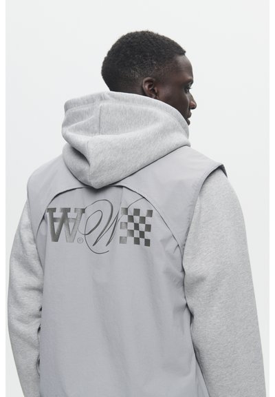 Un sweat à capuche gris avec un gilet de superposition gris. Présente un motif à damier et un logo au dos. Le matériau semble doux et texturé.