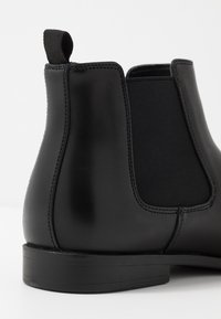 Bottine Chelsea en cuir noir avec panneaux latéraux élastiques, bout arrondi et languette de traction. Texture lisse avec une finition mate. Semelle en caoutchouc plate et basse.