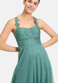 Turkusowa sukienka z koronkowym gorsetem zdobionym kwiatowymi wzorami, regulowanymi ramiączkami i zwiewną spódnicą. Lekki materiał o miękkiej teksturze.