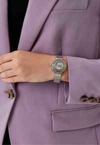 Srebrny siatkowy zegarek na nadgarstek z okrągłą tarczą z koncentrycznymi pierścieniami i kryształowymi akcentami, noszony na ręce przeciwko jasnofioletowej marynarce.