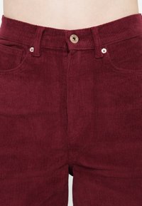 Burgundy kordbársony nadrág, texturált felülettel, elülső zsebekkel, fémből készült gombos derékzárással és látható varrási részletekkel.